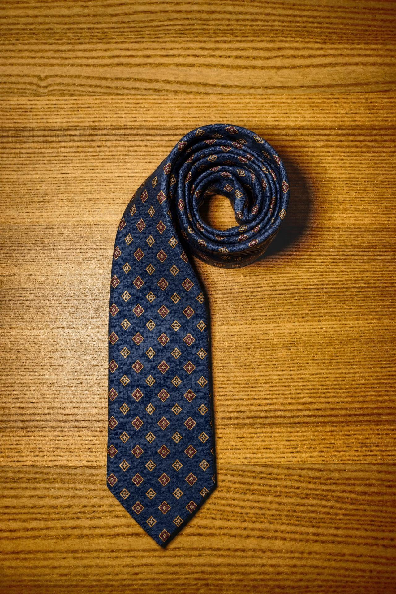 A navy silk foulard tie printed with a small gold and oxblood geometric neat, rolled, on a wooden surface